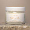 Jar of 'drifted body scrub' on a stone surface with a beige background